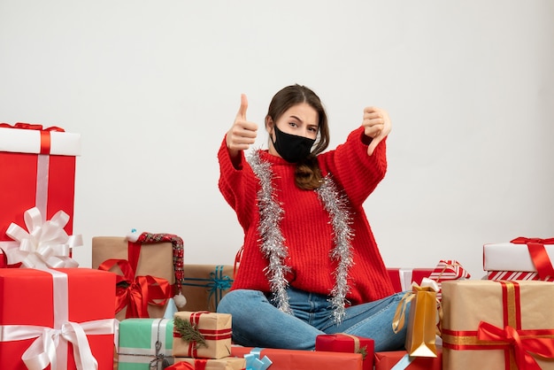 young girl with red sweater making thumb up and thumb down signs sitting around presents with black mask on white