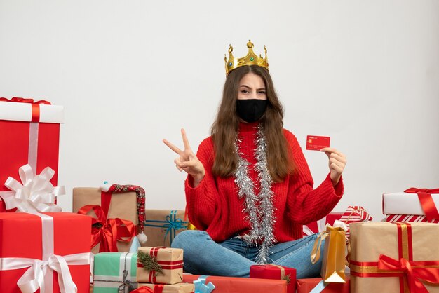 young girl with red sweater making victory sign sitting around presents with black mask on white