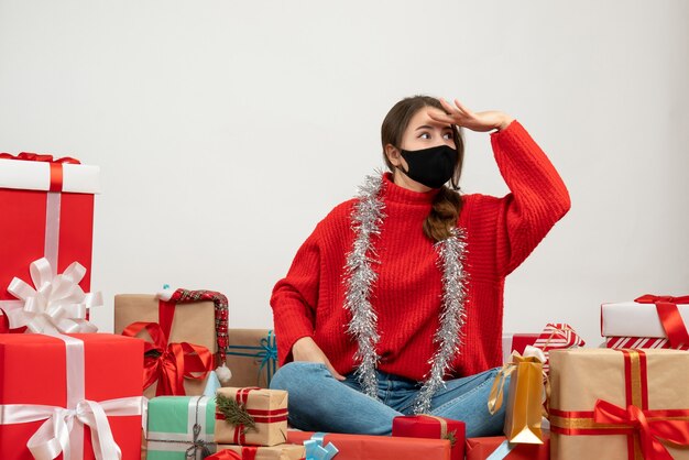 young girl with red sweater observing something sitting around presents with black mask on white