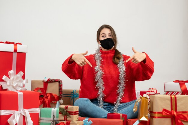 young girl with red sweater pointing with finger herself sitting around presents with black mask on white