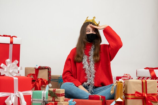 young girl with red sweater putting hand to her forehead sitting around presents with black mask on white