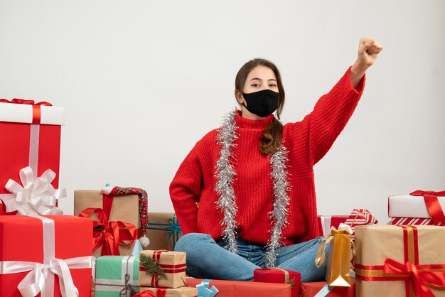 young girl with red sweater raising her hand sitting around presents with black mask on white