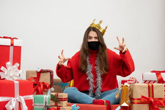 young girl with red sweater showing scary gesture sitting around presents with black mask on white