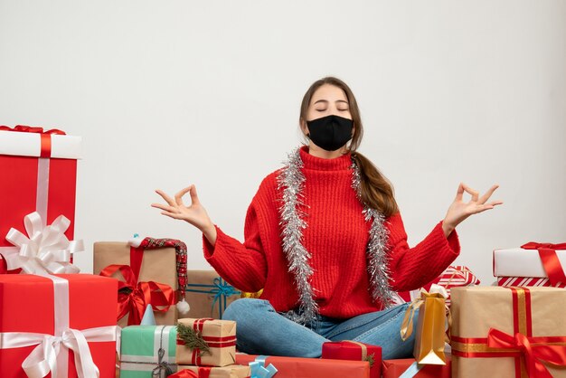 Young girl with red sweater sitting in yoga pose around presents on white