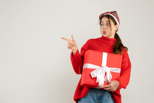 young girl with santa hat holding present pointing with finger something standing on white