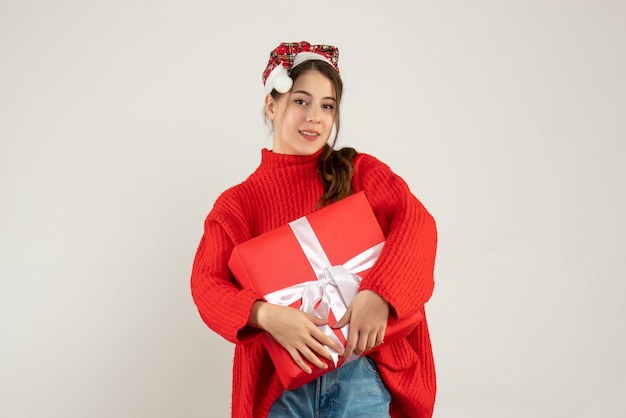 young girl with santa hat holding present with both hands standing on white