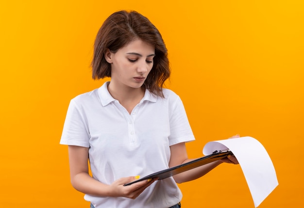 Young girl with short hair wearing polo shirt holding clipboard with blank pages looking at it with serious face