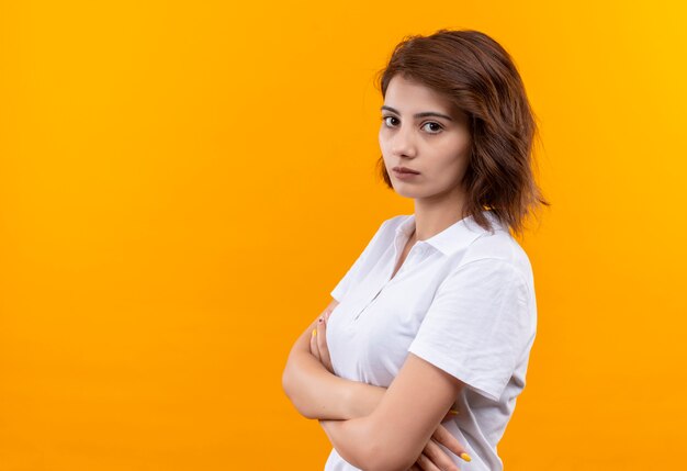 Young girl with short hair wearing polo shirt standing sideways with crossed arms looking at camera with serious face over orange background