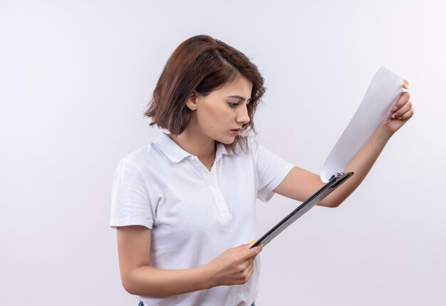 Young girl with short hair wearing white polo shirt holding clipboard looking at blank pages with serious face 