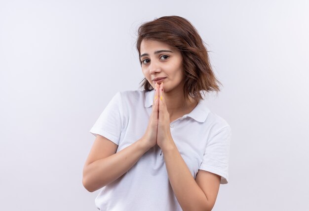 Young girl with short hair wearing white polo shirt holding hands together like praying or begging with hope expression 