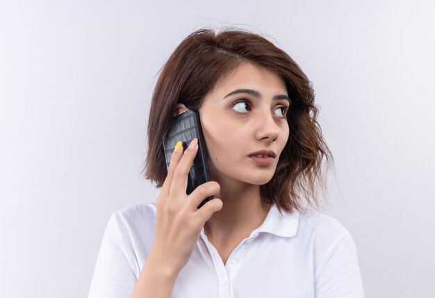 Молодая девушка с короткими волосами в белой рубашке поло, смущенная в сторону, разговаривает по мобильному телефону