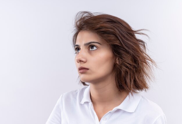 Young girl with short hair wearing white polo shirt looking aside with sad expression on face 