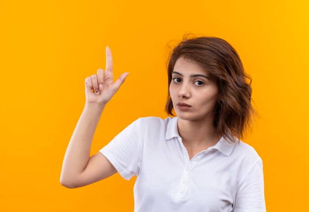 Young girl with short hair wearing white polo shirt looking at camera with serious face pointing with index finger up