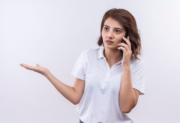 Foto gratuita giovane ragazza con i capelli corti che indossa una polo bianca che sembra confusa e molto ansiosa mentre parla al telefono cellulare