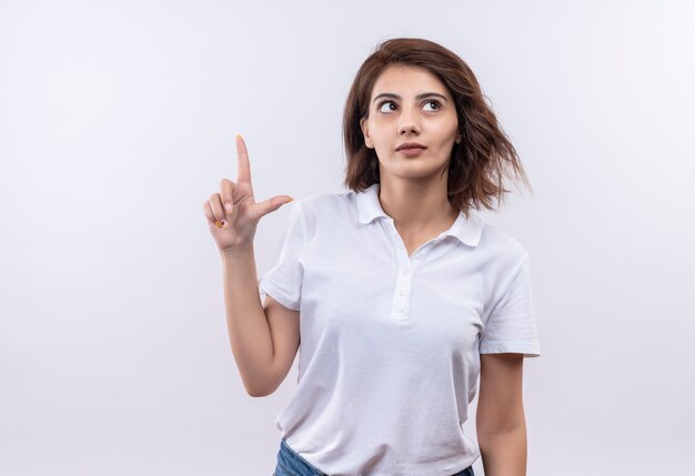 Young girl with short hair wearing white polo shirt looking intrigued pointing up with index finger 