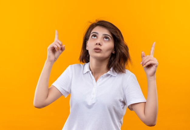 Young girl with short hair wearing white polo shirt looking up confused pointing up with index fingers