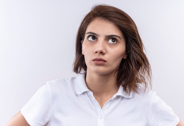 Young girl with short hair wearing white polo shirt looking up with suspicious expression 