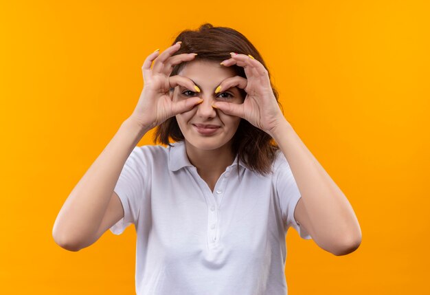 Young girl with short hair wearing white polo shirt making ok signs with fingers like binoculars looking through fingers smiling