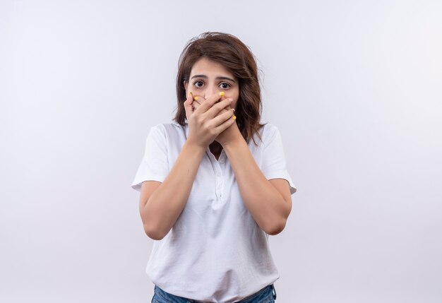Young girl with short hair wearing white polo shirt shocked covering mouth with hands 