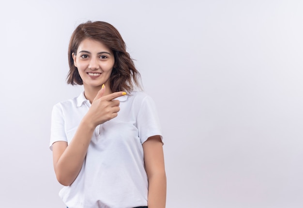 Young girl with short hair wearing white polo shirt smiling positive and happy pointing with finger to the side 