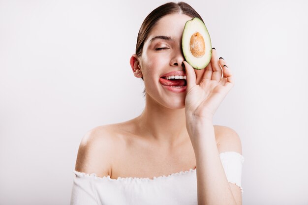 Young girl without makeup in white top licks her lips, posing with tasty and healthy avocado on isolated wall.