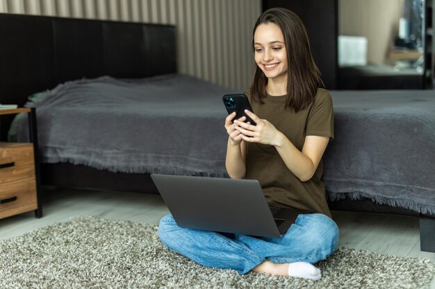 Молодая девушка работает ноутбук, используя смартфон, проверьте уведомление в социальной сети, лежит ковер на полу в доме в помещении