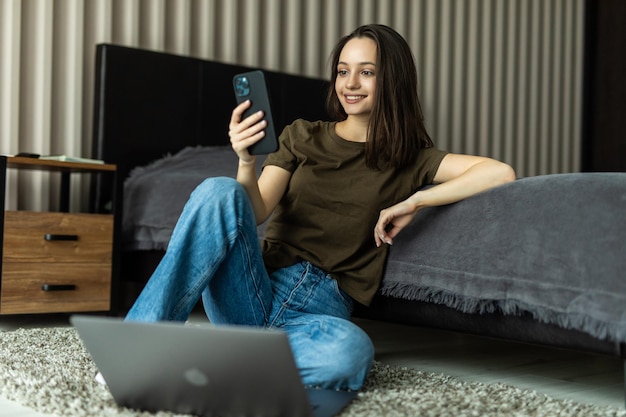 Молодая девушка работает ноутбук, используя смартфон, проверьте уведомление в социальной сети, лежит ковер на полу в доме в помещении