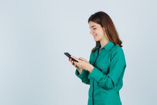 Young girl writing messages on phone in green blouse, black pants and looking cheerful , front view.