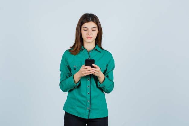 Young girl writing messages on phone in green blouse, black pants and looking focused. front view.