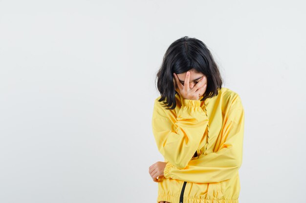 Free photo young girl in yellow bomber jacket covering face with hand, thinking about something and looking pensive