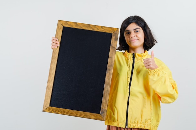 Free photo young girl in yellow bomber jacket holding blackboard, showing thumb up and looking serious