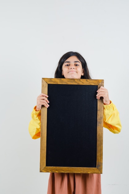 Young girl in yellow bomber jacket holding blackboard with both hands and looking serious