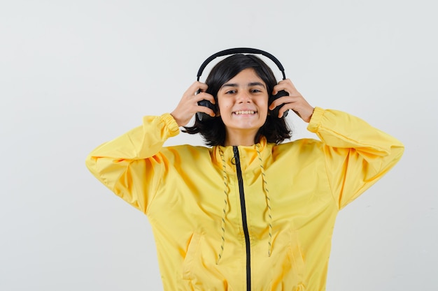 Young girl in yellow bomber jacket holding headphones with both hands and looking happy