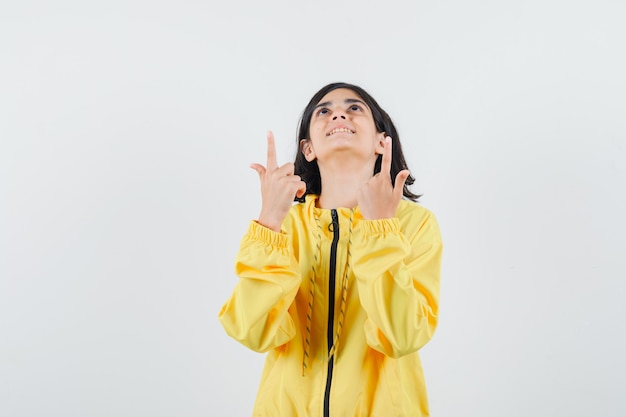 Молодая девушка в желтой куртке-бомбардировке указывает вверх указательными пальцами и выглядит счастливой
