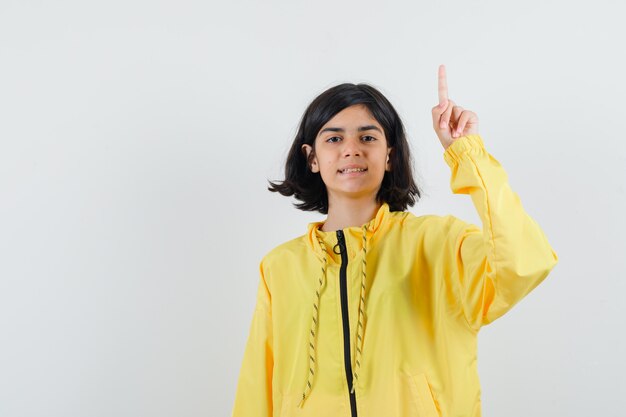 Young girl in yellow bomber jacket raising index finger in eureka gesture and looking happy