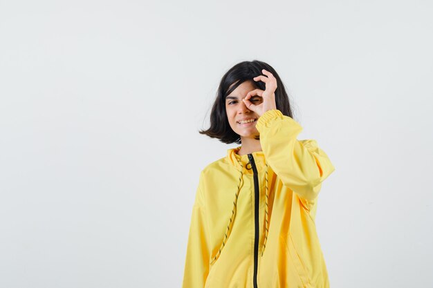 Young girl in yellow bomber jacket showing okay sign on eye and looking happy