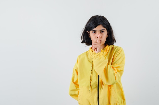 Young girl in yellow bomber jacket showing silence gesture and looking serious