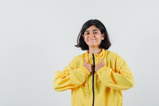 Молодая девушка в желтой куртке-бомбардировщике показывает палец вверх обеими руками и выглядит счастливой