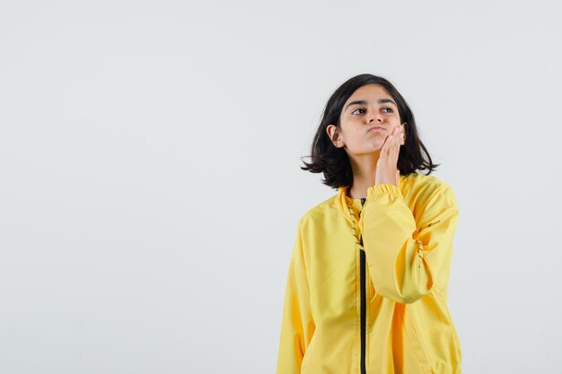 Young girl in yellow bomber jacket standing in thinking pose, putting hand on cheek and looking pensive