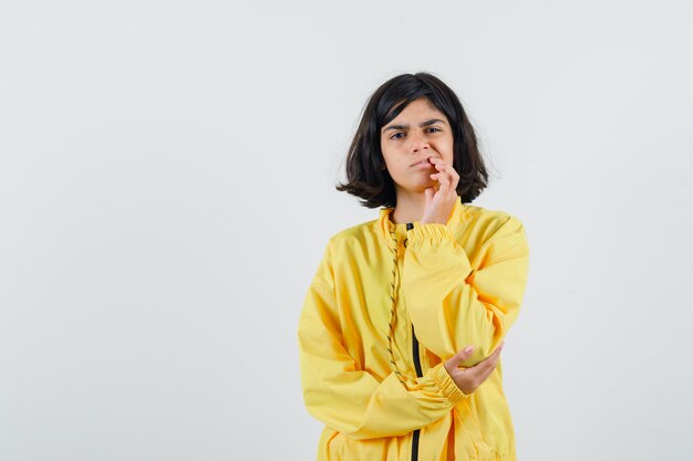 Young girl in yellow bomber jacket standing in thinking pose, putting hand on mouth and looking pensive