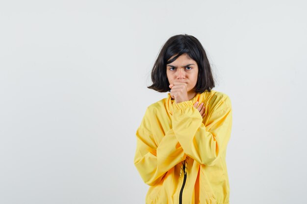 Молодая девушка в желтой куртке-бомбардировщике стоит в позе размышлений, положив руку на рот и задумчиво