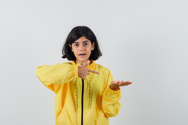 Young girl in yellow bomber jacket stretching hand as holding something and pointing to it and looking surprised