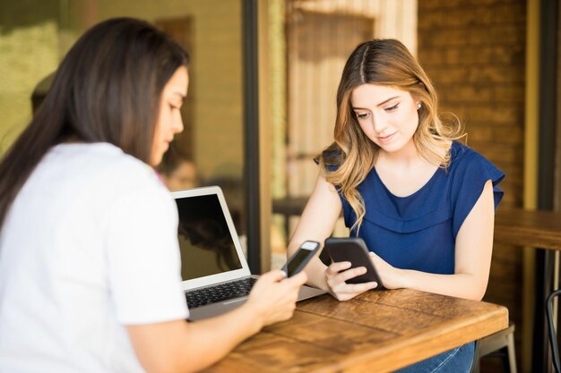 テーブルの上にラップトップで自分の携帯電話を使用してカフェでぶらぶらしている若い女の子