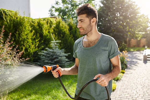 Young good-looking bearded caucasian man with stylish hairstyle in blue t-shirt concentrated watering garden with hose.