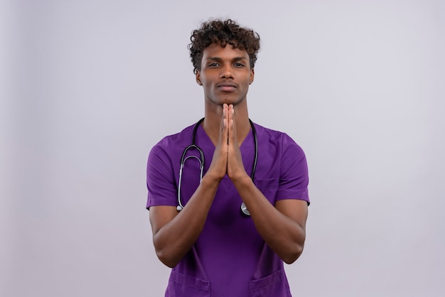 A young good-looking dark-skinned male doctor with curly hair wearing violet uniform with stethoscope holding hand together 