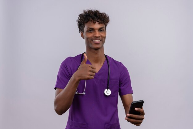 OKのジェスチャーを見せながら彼のスマートフォンを保持している聴診器で紫の制服を着た巻き毛の若い見栄えの良い暗い肌の男性医師