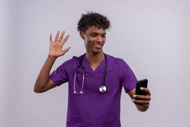 スマートフォンを振っている手を見て聴診器で紫の制服を着た巻き毛の若い見栄えの良い暗い肌の男性医師