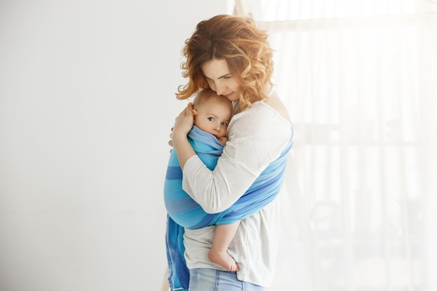 Young good-looking mother snuggles and calming her newborn son who feeling scared after long sleep interrupted by loud sounds from street. Scene of protection and love.