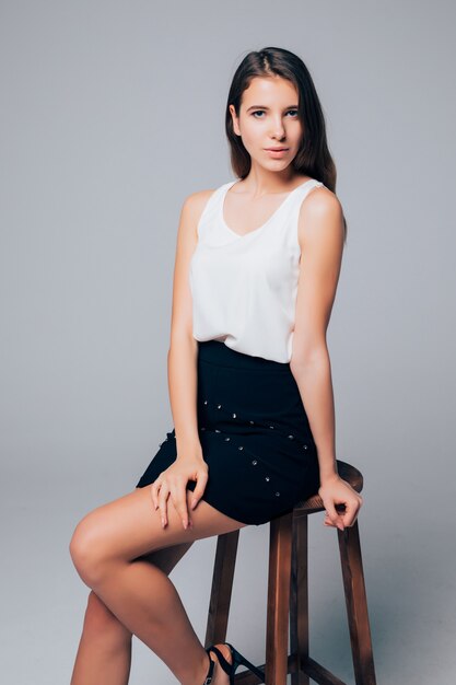 Young gorgeous brunette in short black skirt sit on high wood chair isolated on white