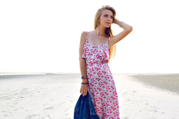 Молодая великолепная женщина с удивительными длинными светлыми волосами позирует на закате на пляже, в стильном винтажном платье. Романтическое настроение.